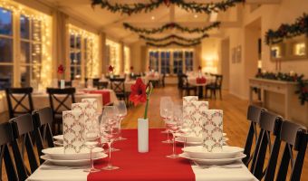 Festlich gedeckte Weihnachtstafel in einem Restaurant mit rotem Tischläufer, elegantem Geschirr, Weingläsern, weihnachtlichen Servietten und stimmungsvoller Weihnachtsdekoration mit Tannengirlanden und Lichterketten.