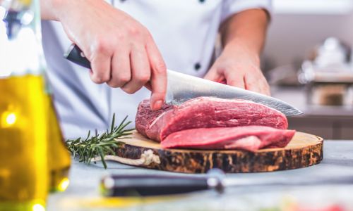 Person in weißer Kochjacke schneidet rohes Rindfleisch auf einem Holzbrett, daneben liegen Rosmarinzweige.