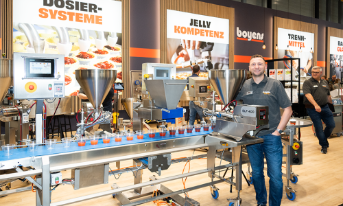 Ein Mann steht hinter einer Portioniermaschine, die neben einem Fließband montiert wurde, um Lebensmittelzutaten in Bechern auf dem Fließband zu portionieren.