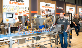 Ein Mann steht hinter einer Portioniermaschine, die neben einem Fließband montiert wurde, um Lebensmittelzutaten in Bechern auf dem Fließband zu portionieren.