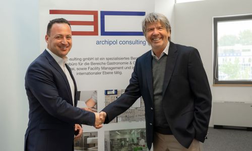 Die beiden geschäftsführenden Gesellschafter schütteln sich die Hand vor einem Aufsteller von Archipol Consulting.