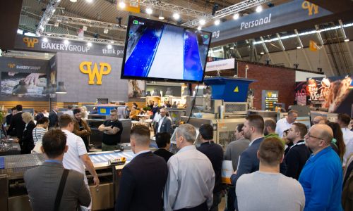Zahlreiche Südback-Besucher stehen vor dem Messestand von WP und sehen sich Produktionsmaschinen für das Bäckerhandwerk an. Die Vorführung der Maschinen wird auch auf einem Bildschirm angezeigt.