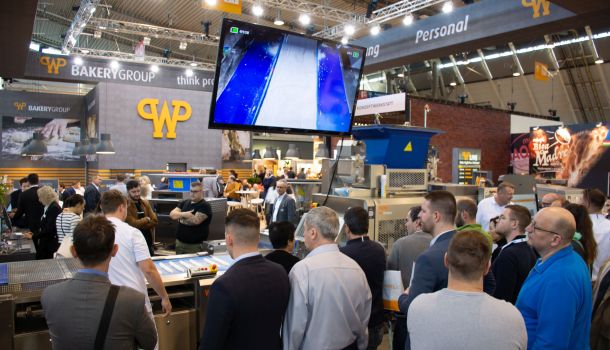Zahlreiche Südback-Besucher stehen vor dem Messestand von WP und sehen sich Produktionsmaschinen für das Bäckerhandwerk an. Die Vorführung der Maschinen wird auch auf einem Bildschirm angezeigt.