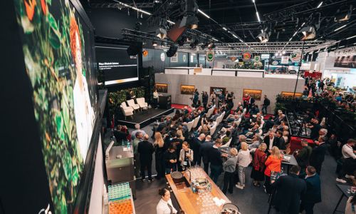 In einer Messehalle sitzen viele Menschen auf Stühlen vor einer Bühne, auf der leere Stühle stehen.