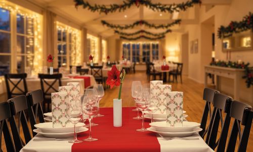 Festlich gedeckte Weihnachtstafel in einem Restaurant mit rotem Tischläufer, elegantem Geschirr, Weingläsern, weihnachtlichen Servietten und stimmungsvoller Weihnachtsdekoration mit Tannengirlanden und Lichterketten.