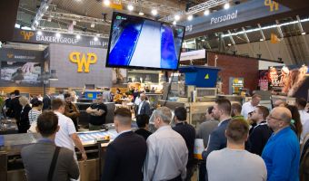 Zahlreiche Südback-Besucher stehen vor dem Messestand von WP und sehen sich Produktionsmaschinen für das Bäckerhandwerk an. Die Vorführung der Maschinen wird auch auf einem Bildschirm angezeigt.